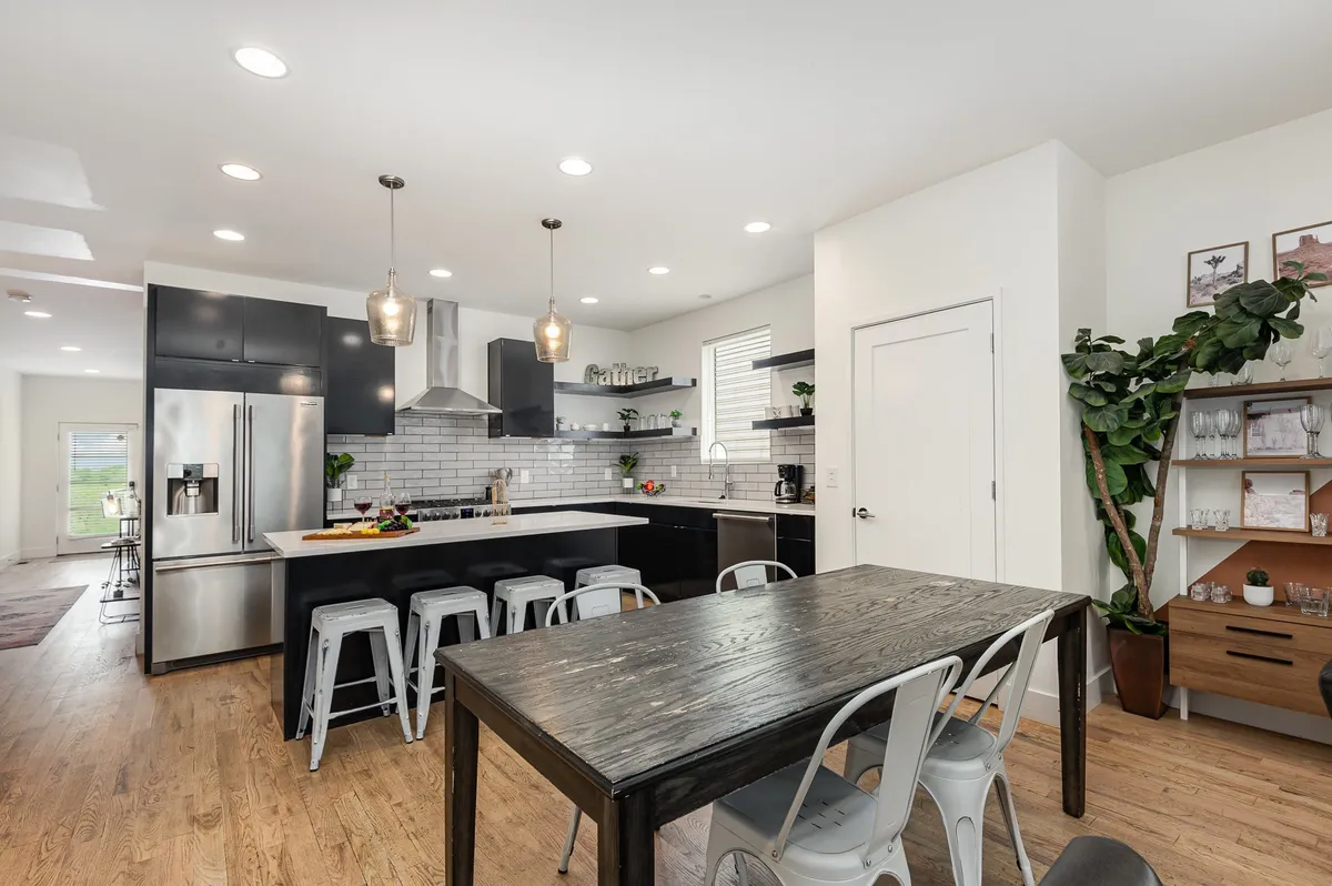 A bright, modern kitchen and dining area featuring a large island with seating, a spacious dining table, black cabinetry, stainless steel appliances, and green plants as accents. The open-plan design encourages gatherings for meals and celebrations.