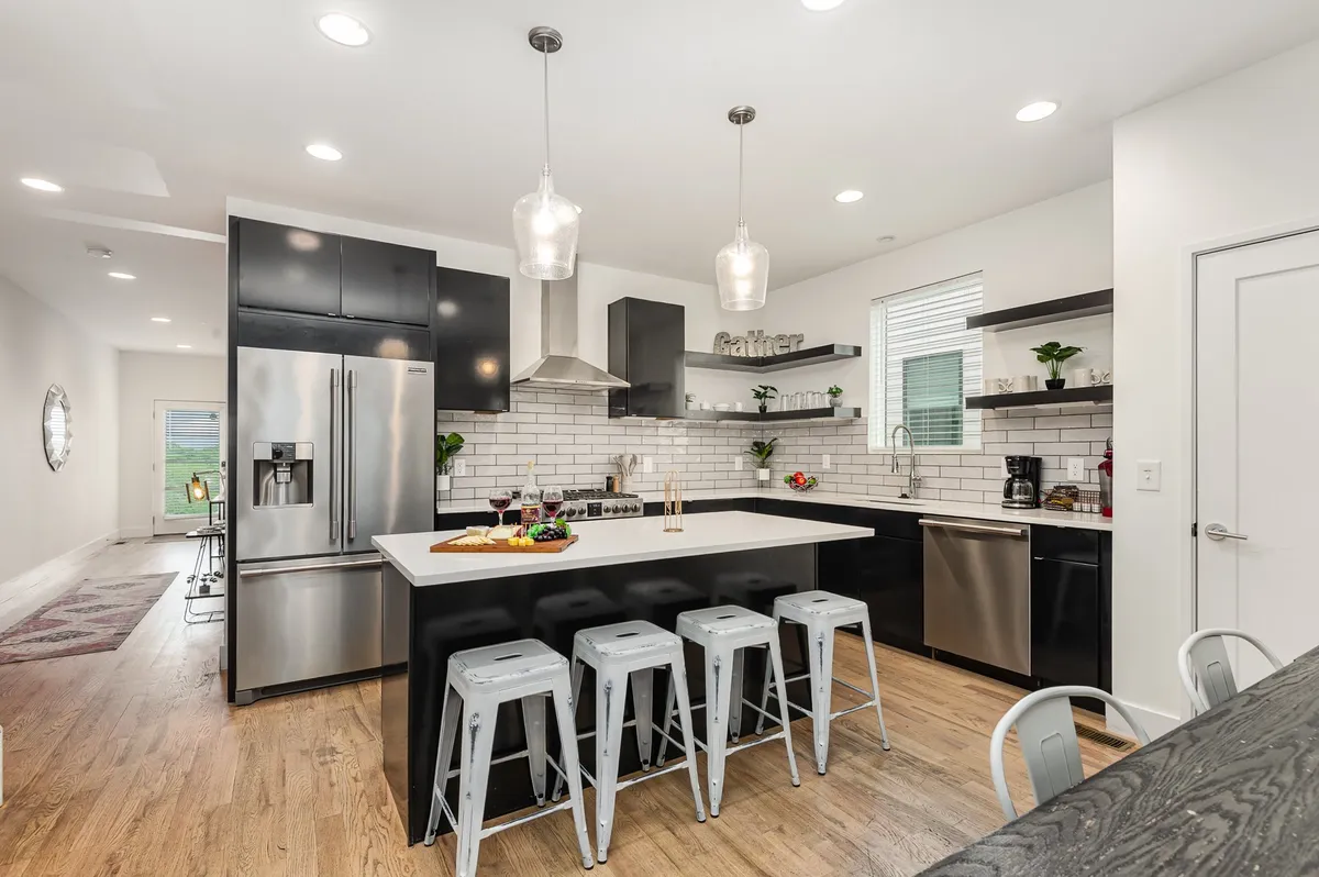 A warm and welcoming kitchen scene featuring a spacious kitchen island with bar stools, modern stainless steel appliances, and open shelving with lush green plants. The island provides ample space for meal preparation and casual dining, while the overall design radiates a homey, inviting atmosphere perfect for gatherings.