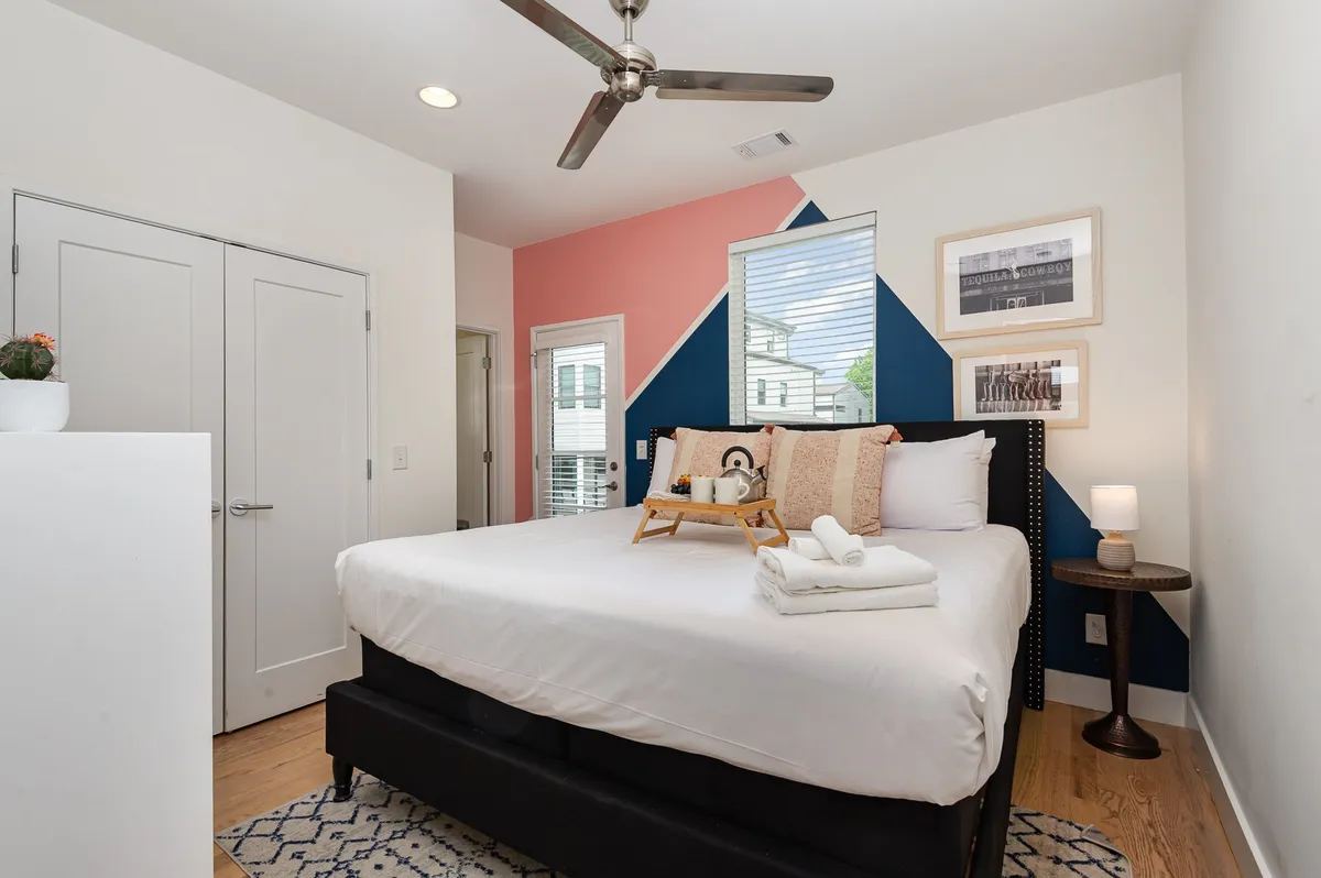 A stylish and inviting bedroom featuring a neatly made bed with colorful accent pillows and towels, a bold accent wall, and framed artwork. The space exudes comfort and a sense of tranquility, ideal for restful nights or peaceful mornings.