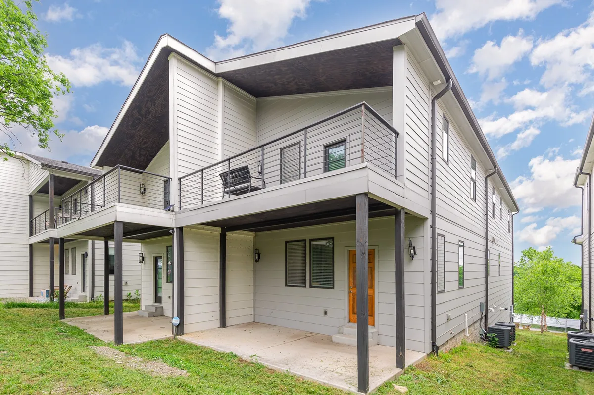 The image showcases the dual-home exterior with a view of the balcony, front door, and the green lawn in front. The design clearly demonstrates the presence of two adjacent, fully independent residences situated side-by-side, with private entrances and a shared driveway area.