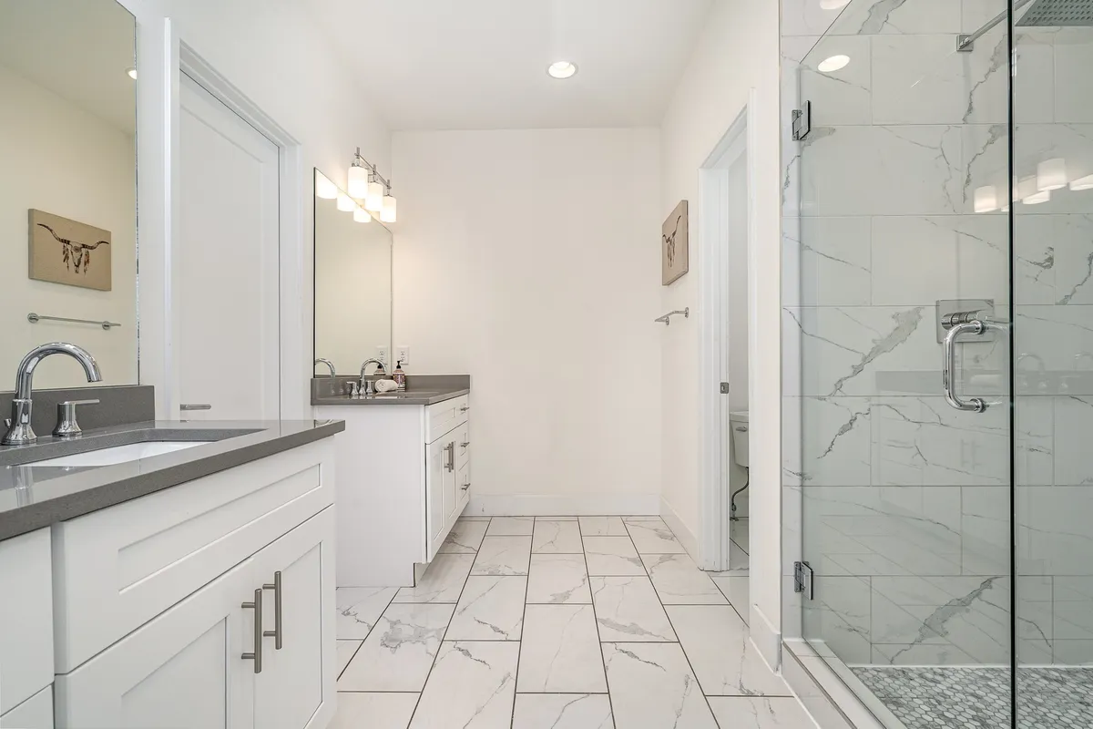 A contemporary bathroom with a double vanity sink, a glass shower enclosure, a large mirror, and modern artwork. The serene and spacious layout is ideal for self-care routines and provides privacy for guests.
